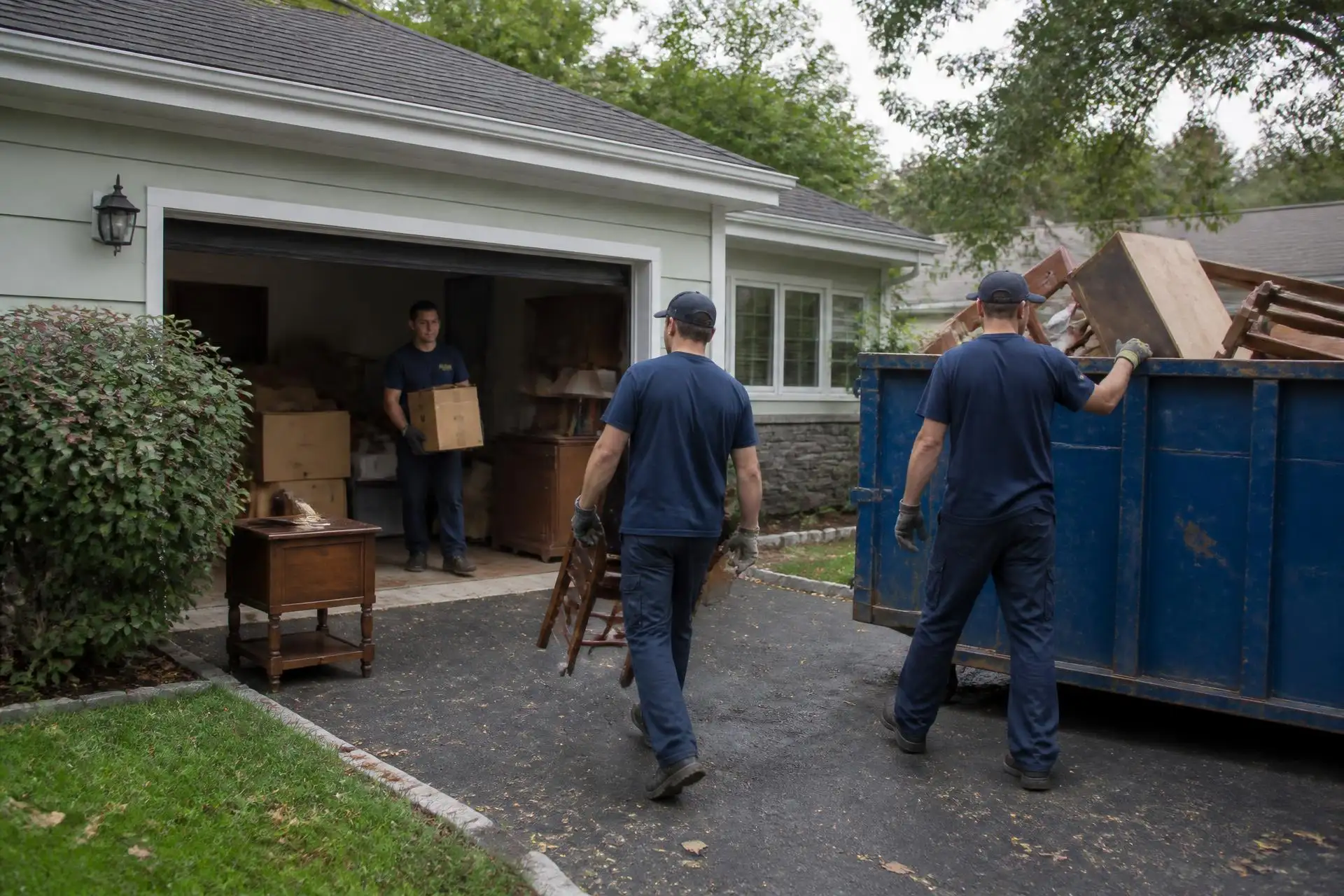 Workers carrying boxes and furniture out of a garage into a driveway container during an estate cleanout